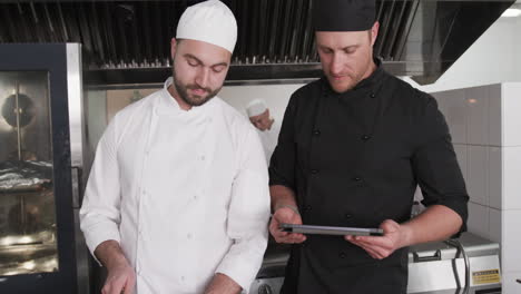 Focused-cucasian-male-chef-instructing-trainee-male-chef-using-tablet-in-kitchen,-slow-motion