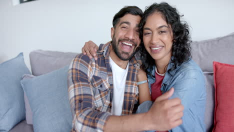 Portrait-of-happy-biracial-couple-sitting-on-sofa-embracing-and-smiling-at-home,-in-slow-motion