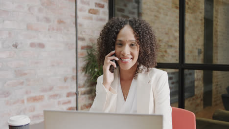 Portrait-of-happy-biracial-businesswoman-using-laptop-and-talking-on-smartphone,-in-slow-motion