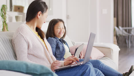 Video-of-happy-asian-mother-and-daughter-sitting-on-sofa-with-laptop-and-tablet