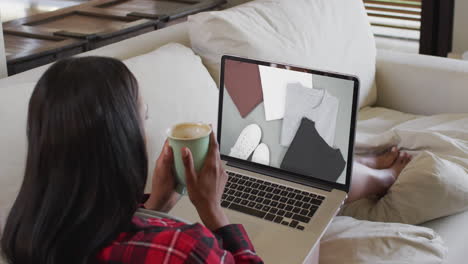 Biracial-woman-using-laptop-on-couch-at-home-online-shopping-for-clothing,-slow-motion