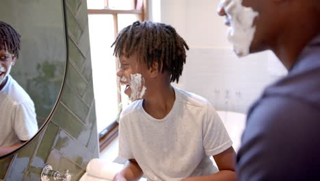 Happy-african-american-father-and-son-applying-shaving-foam-on-face-in-bathroom-at-home,-slow-motion