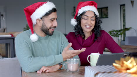 Happy-biracial-couple-wearing-santa-claus-hats-using-tablet-for-video-call-at-home,-in-slow-motion