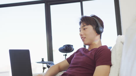 Glücklicher-Asiatischer-Männlicher-Teenager,-Der-Kopfhörer-Trägt-Und-Im-Wohnzimmer-Einen-Laptop-Benutzt
