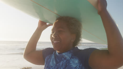 Glückliche-ältere-Afroamerikanische-Frau,-Die-In-Zeitlupe-Am-Strand-Spazieren-Geht-Und-Ein-Surfbrett-Hält