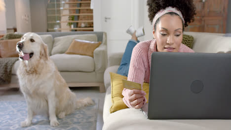 Happy-biracial-woman-with-golden-retriever-dog-using-laptop-and-credit-card-at-home,-slow-motion