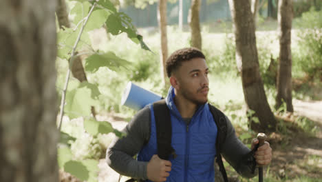 African-american-man-wearing-backpack-and-hiking-with-trekking-poles-in-forest,-slow-motion