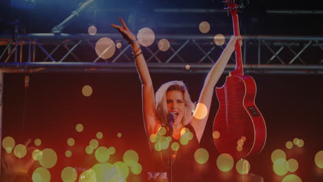 Animation-of-glowing-spots-falling-over-caucasian-female-singer-holding-a-guitar-singing-at-concert