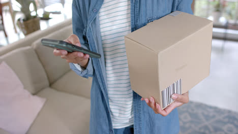 Midsection-of-biracial-woman-holding-parcel-and-using-smartphone-at-home,-slow-motion