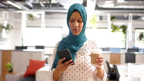 Biracial-casual-businesswoman-in-hijab-drinking-coffee-and-using-smartphone-in-office,-slow-motion