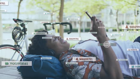 Animation-of-social-media-icons-over-african-american-man-using-smartphone-lying-on-bench-on-street