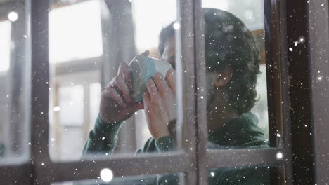 Animation-of-window-and-snow-falling-over-caucasian-man-drinking-coffee-at-christmas