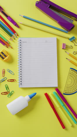 Vertical-video-of-notebook,-coloured-pens,-pencils-and-school-stationery-on-yellow-background