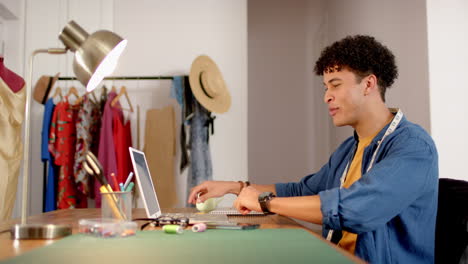 Biracial-male-fashion-designer-sketching-design-at-desk-using-laptop-in-studio,-slow-motion