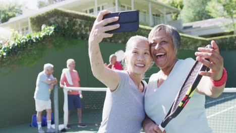 Two-happy-diverse-senior-women-taking-selfie-at-sunny-outdoor-tennis-court-with-friends,-slow-motion