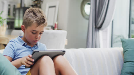 Happy-caucasian-boy-sitting-on-sofa-and-using-tablet,-copy-space,-slow-motion