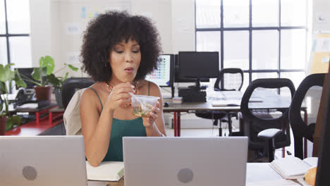 Happy-biracial-casual-businesswoman-sitting-at-desk,-eating-and-looking-at-laptop,-slow-motion