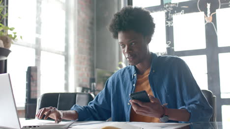 African-american-casual-businessman-sitting-at-desk-using-smartphone-and-laptop-at-home,-slow-motion