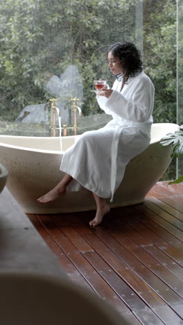Vertical-video-of-biracial-woman-sitting-on-bathtub-drinking-tea-at-home,-slow-motion