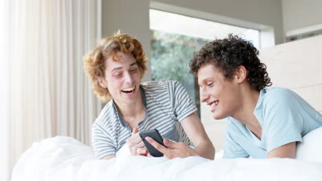 Happy-diverse-gay-male-couple-lying-on-bed-and-using-smartphone-at-home,-slow-motion