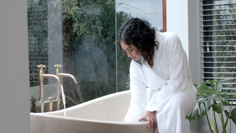 Biracial-woman-wearing-bathrobe-sitting-on-bathtub-running-bath-in-bathroom-at-home,-slow-motion