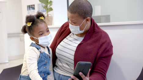 African-american-mother-and-daughter-wearing-face-masks-using-smartphone-in-hospital,-slow-motion