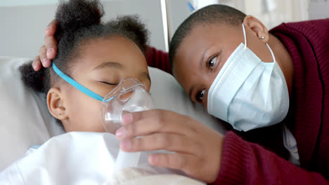 African-american-woman-with-face-mask-stroking-daughter-with-oxygen-mask-in-hospital,-slow-motion