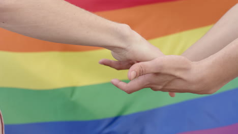 Midsection-of-diverse-gay-male-couple-holding-hands-over-rainbow-lgbt-flag,-slow-motion