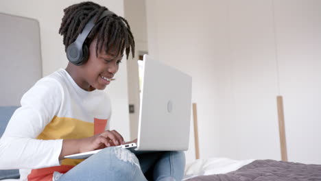 Happy-african-american-boy-wearing-headphones-sitting-on-bed-using-laptop-at-home,-slow-motion