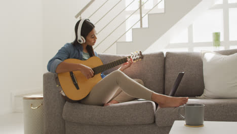Video-of-happy-biracial-woman-in-headphones-sitting-on-sofa-and-playing-guitar