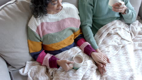 Happy-biracial-couple-sitting-on-sofa-and-drinking-coffee-under-blanket-at-home,-in-slow-motion