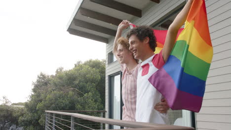 Happy-diverse-gay-male-couple-holding-rainbow-lgbt-flag-on-balcony-at-home,-slow-motion
