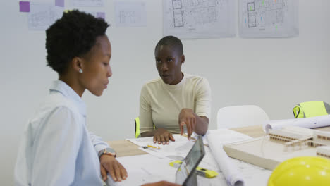 Zwei-Afroamerikanische-Architektinnen-Diskutieren-Mit-Bauplänen-Und-Verwenden-Ein-Tablet-Im-Büro