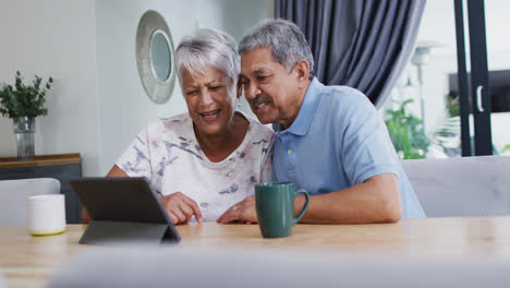 Happy-senior-biracial-couple-laughing-and-using-tablet