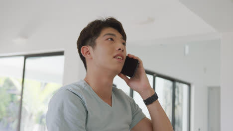 Asian-male-teenager-talking-on-smartphone-and-smiling-in-living-room