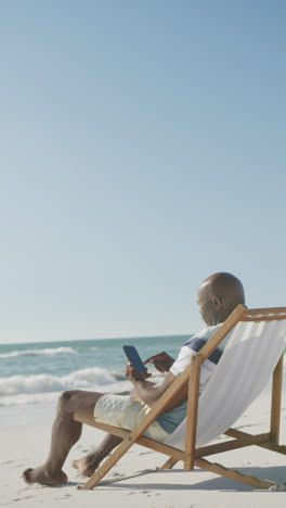 Vertical-video-of-senior-african-american-man-using-smartphone-on-deckchair-at-beach,-in-slow-motion