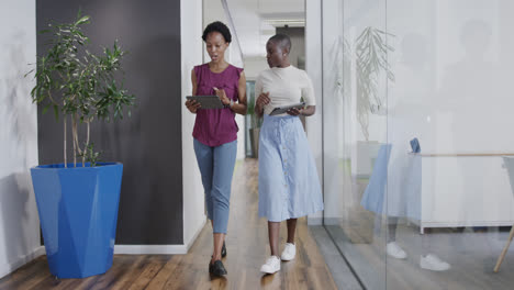 Happy-african-american-businesswomen-with-tablets-talking,-walking-in-office,-in-slow-motion