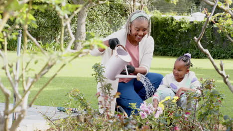 Happy-african-american-grandmother-and-granddaughter-watering-plants-in-sunny-garden,-slow-motion
