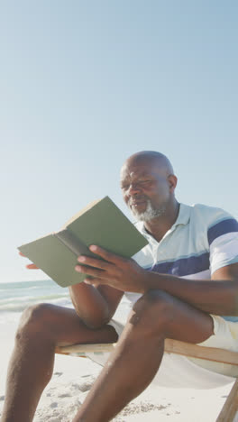 Vertical-video-of-senior-african-american-man-reading-book-on-deckchair-at-beach,-in-slow-motion