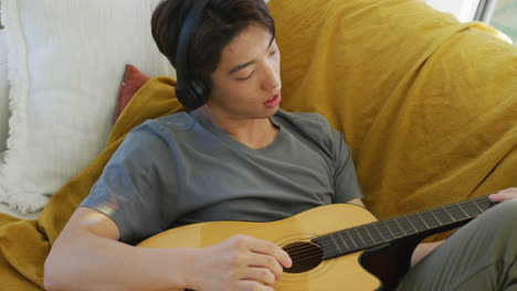 Asian-boy-wearing-headphones-playing-guitar-lying-on-bean-bag-at-home