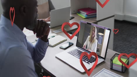 Animation-of-red-heart-icons-falling-over-african-american-man-having-video-call-on-laptop-at-office