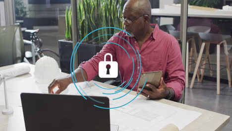 Animation-of-security-padlock-icon-over-african-american-man-working-on-his-plan-at-office