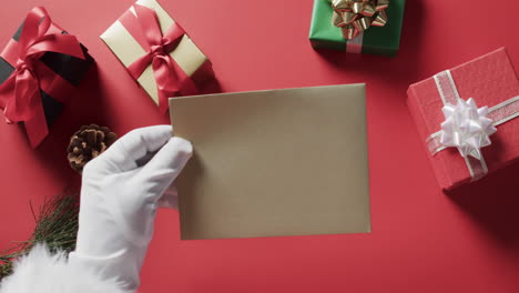Video-of-santa-claus-holding-brown-envelope-with-copy-space-and-christmas-presents-on-red-background