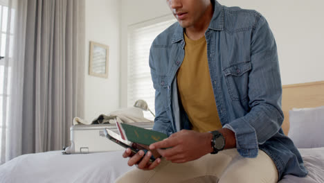 Biracial-man-using-smartphone-and-checking-ticket-and-passport-in-bright-bedroom
