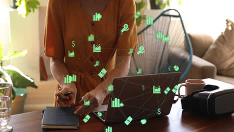 Animation-of-network-of-green-business-icons-over-woman-using-laptop-writing-in-notebook