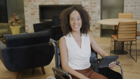 Portrait-of-happy-biracial-businesswoman-in-wheelchair-using-tablet-in-office,-in-slow-motion