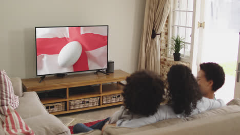 Biracial-family-watching-tv-with-rugby-ball-on-flag-of-england-on-screen