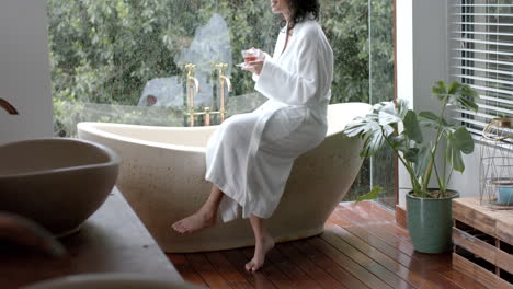 Biracial-woman-wearing-bathrobe-sitting-on-bathtub-drinking-tea-in-bathroom-at-home,-slow-motion