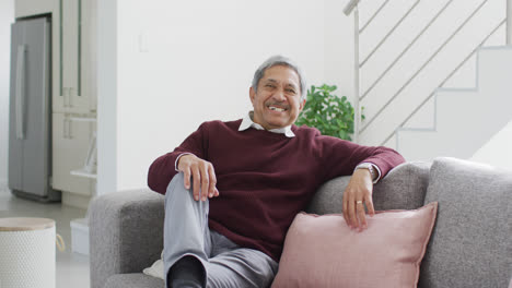 Videoporträt-Eines-älteren-Mannes-Mit-Gemischter-Abstammung,-Der-Im-Wohnzimmer-Auf-Der-Couch-Sitzt-Und-In-Die-Kamera-Lächelt