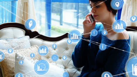 Animation-of-connected-icons-over-caucasian-woman-sitting-on-couch-and-talking-on-cellphone